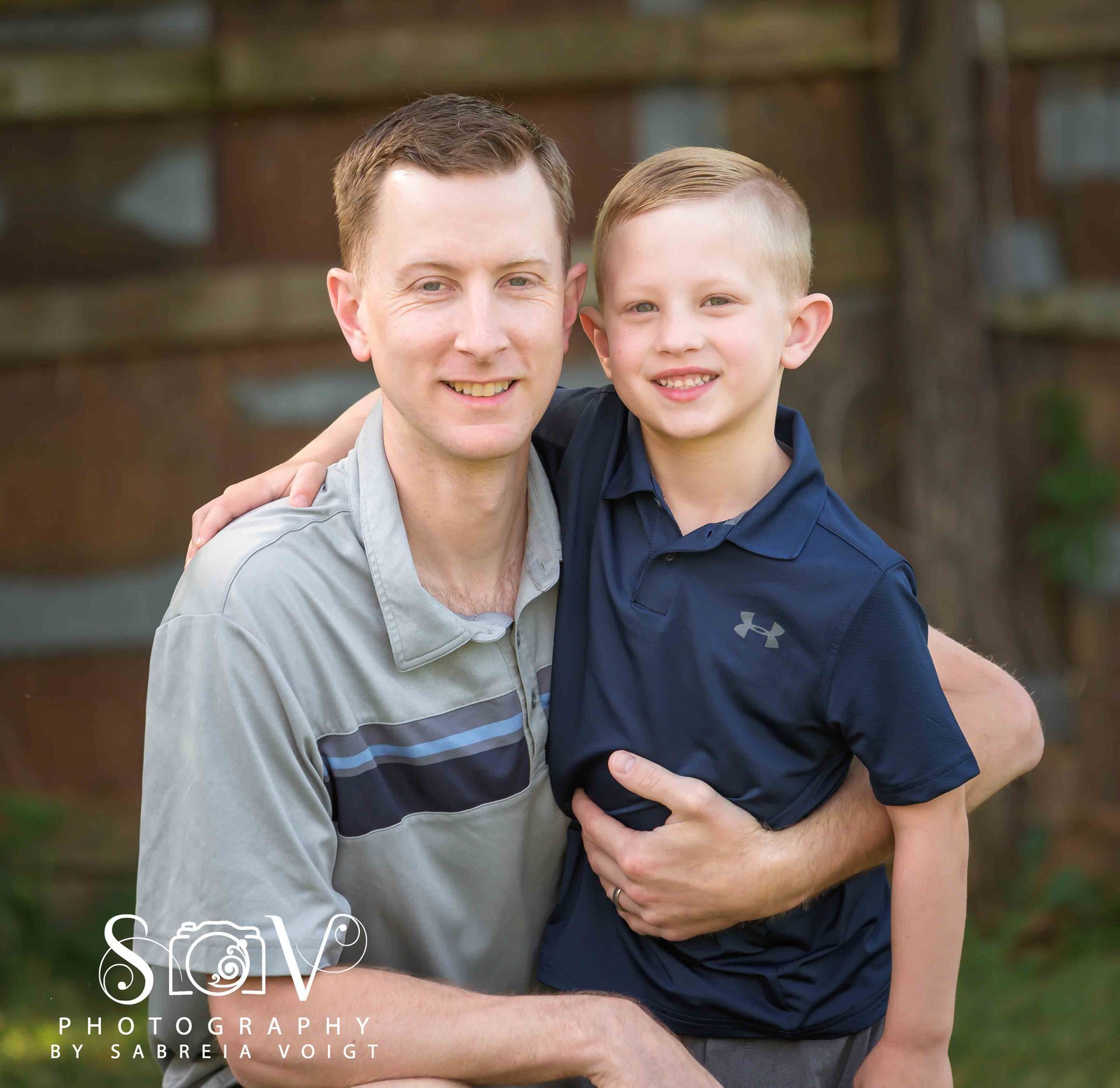 Man with arm around a boy, smiling. They are in front of a wooden fence, outdoors.