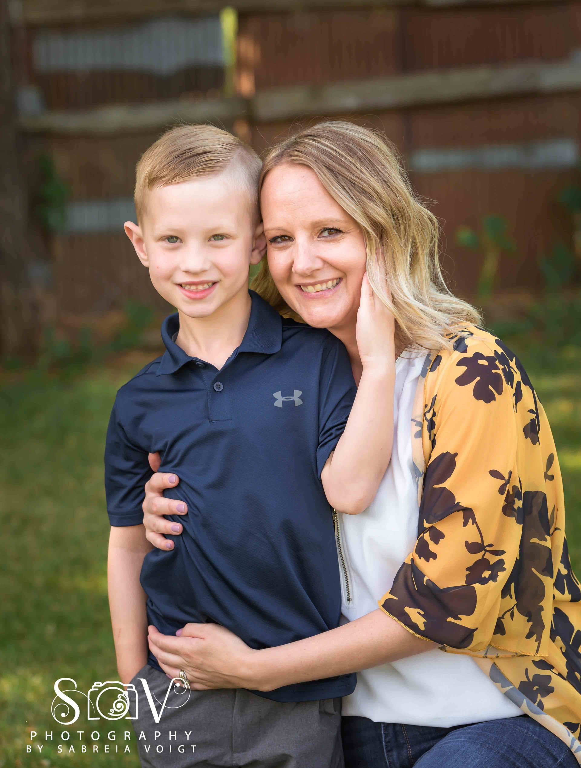 Woman hugs child, smiling. Outdoor setting. Woman wears yellow floral top, child wears blue polo.