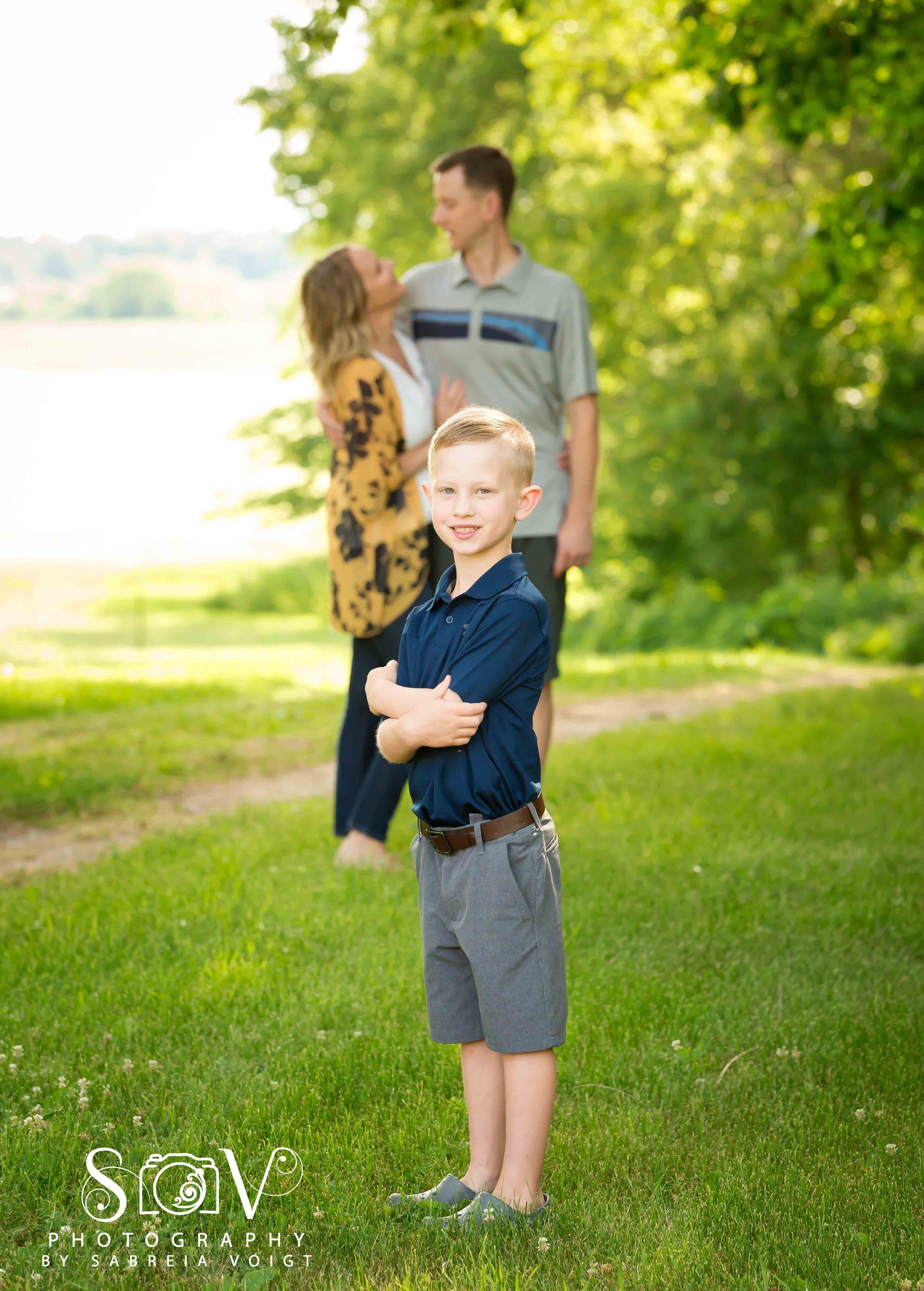 Boy in front with crossed arms, parents embrace in background, park setting.