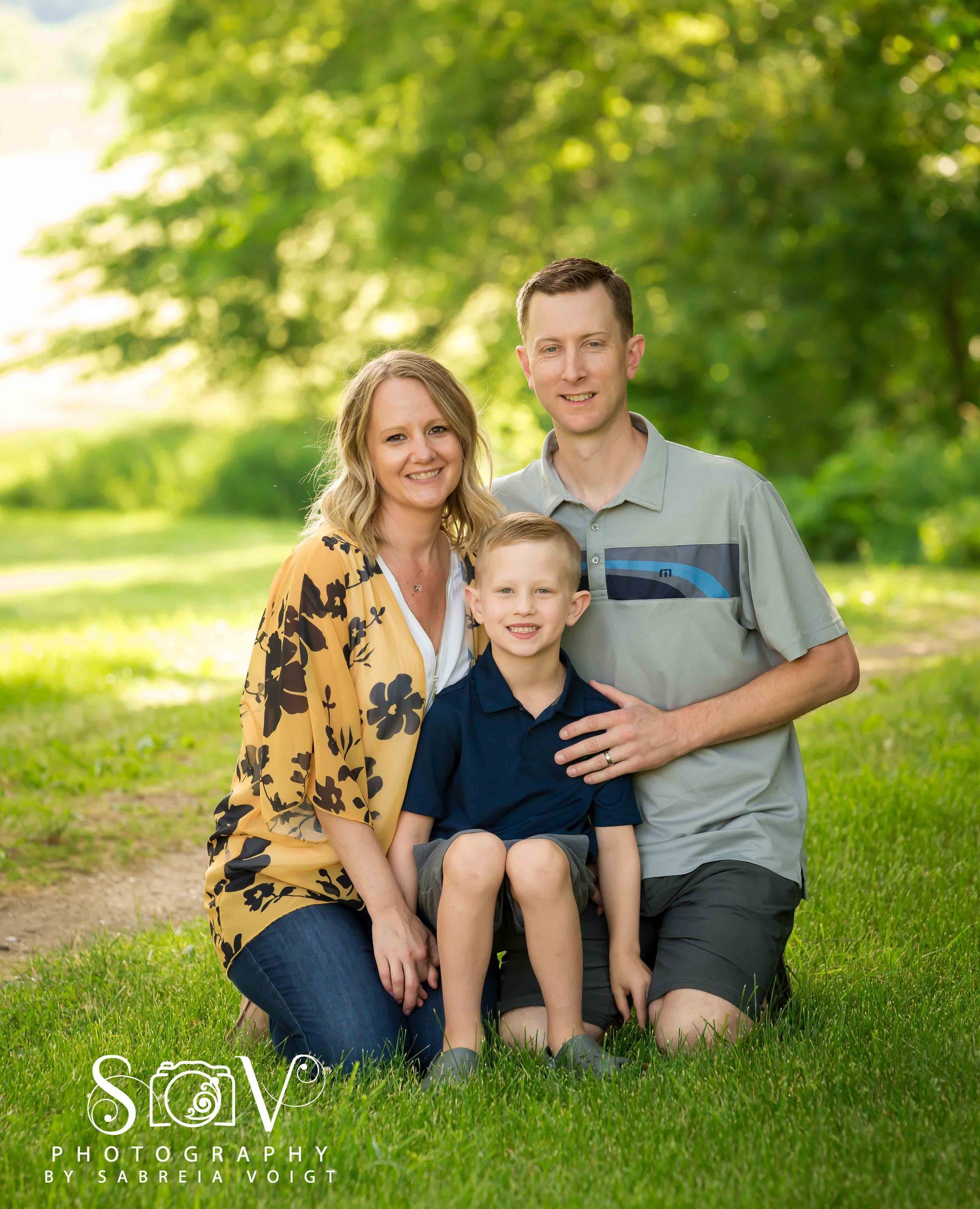 Family kneeling in grass, posing for photo: woman in yellow top, man in grey shirt, child in blue shirt.