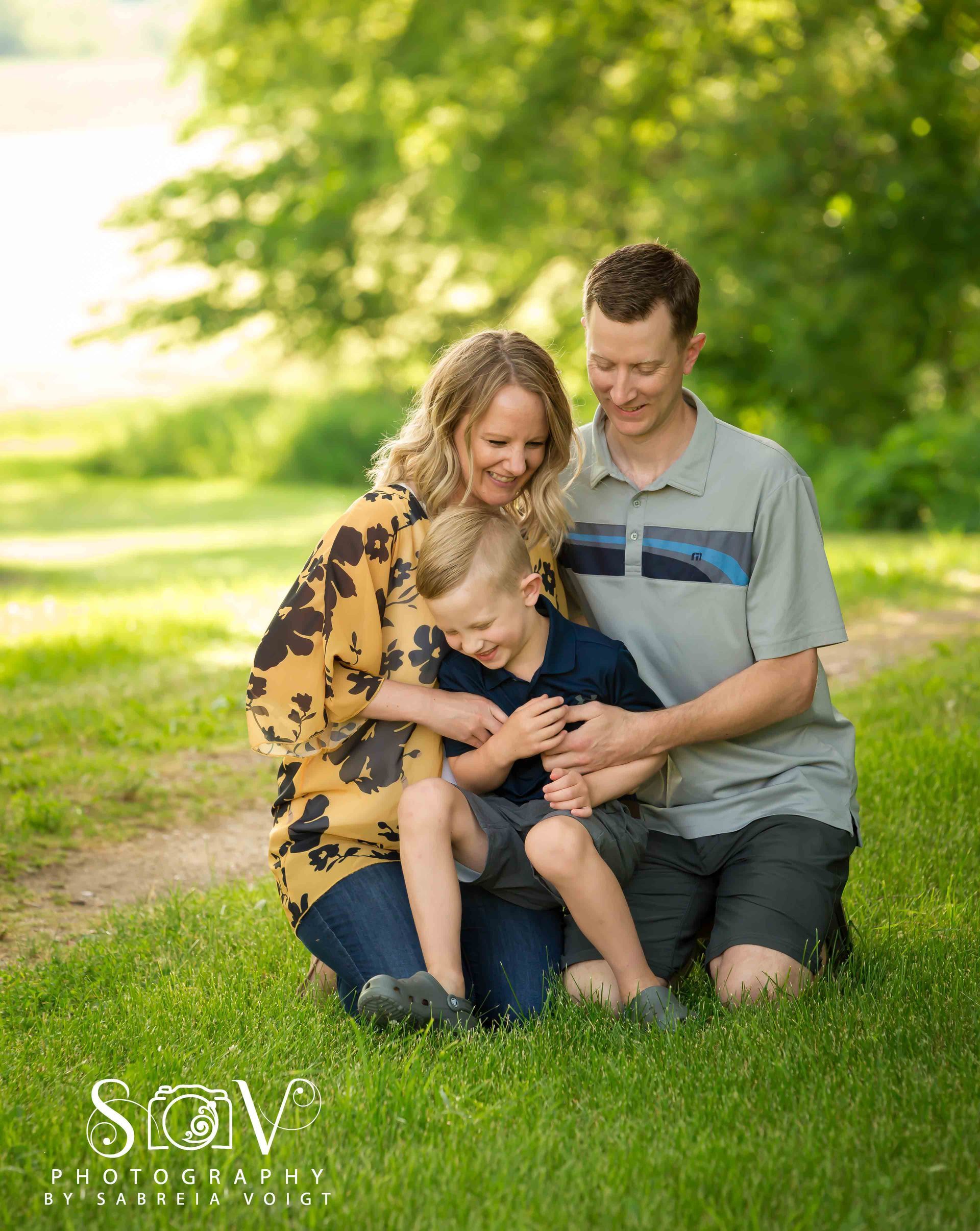 Family kneels together in grassy field. Parents embrace child, smiling. Green trees in background.