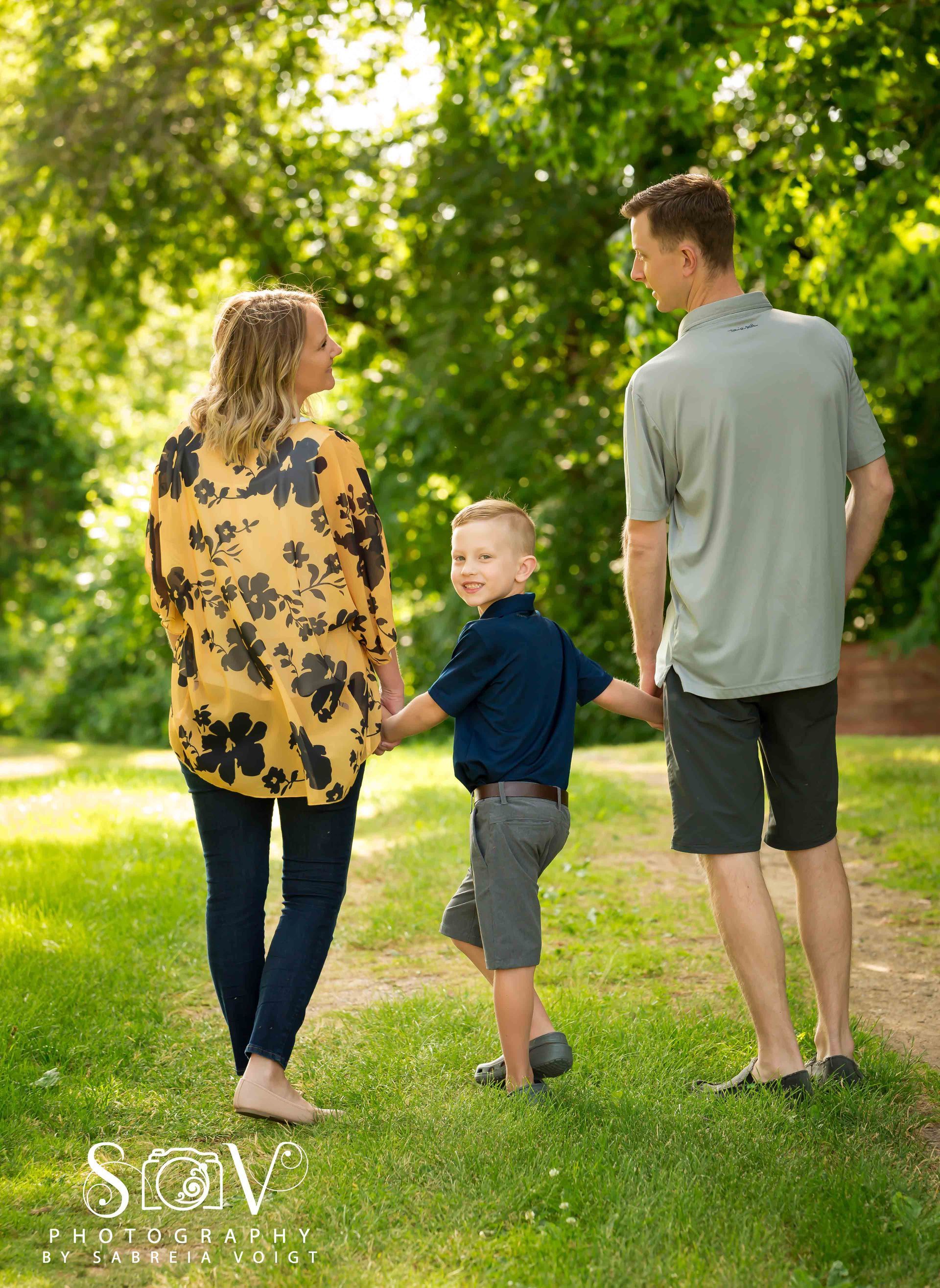 Family, walking in park, holding hands. Mom in floral shirt, son in blue shirt, dad in gray shirt. Sunny day.