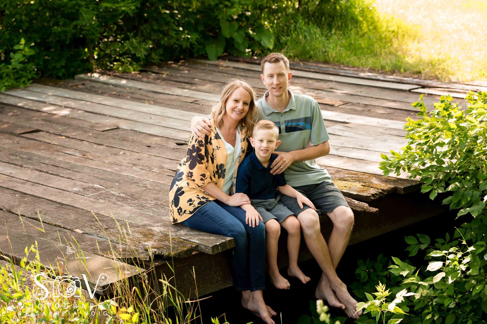 Family of three sits barefoot on a wooden bridge, smiling. Sunlight filters through trees, and foliage surrounds.