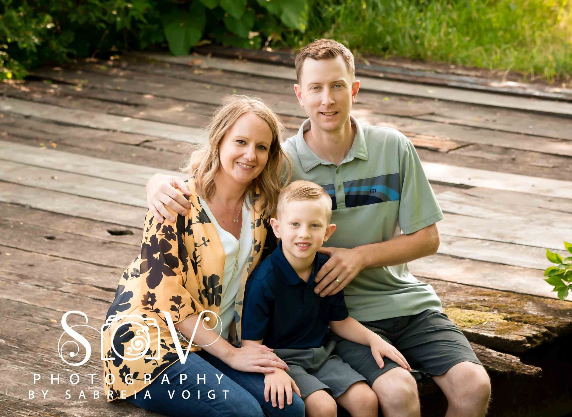 Family of three sitting on a wooden bridge, smiling. Woman in floral top, man in striped shirt, child in blue.