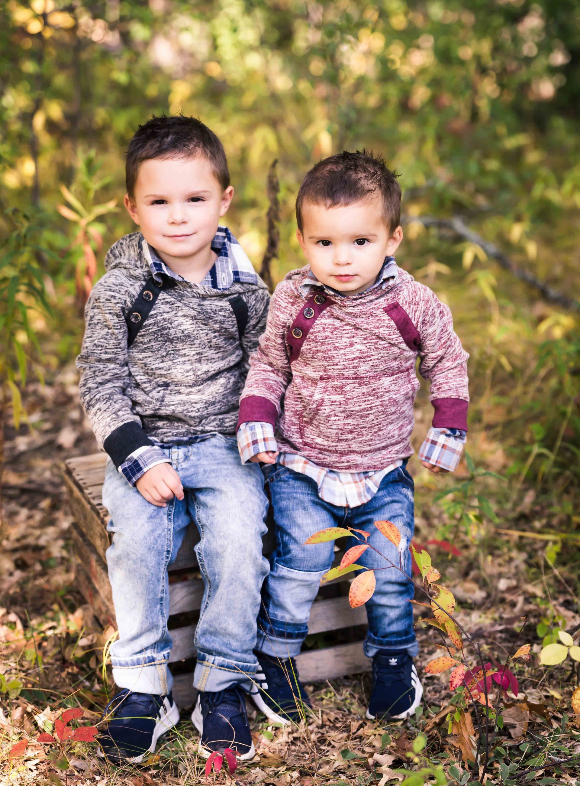 Two boys sitting outdoors on a wooden box. Both wear hoodies and jeans. One looks at the camera. Autumn setting.