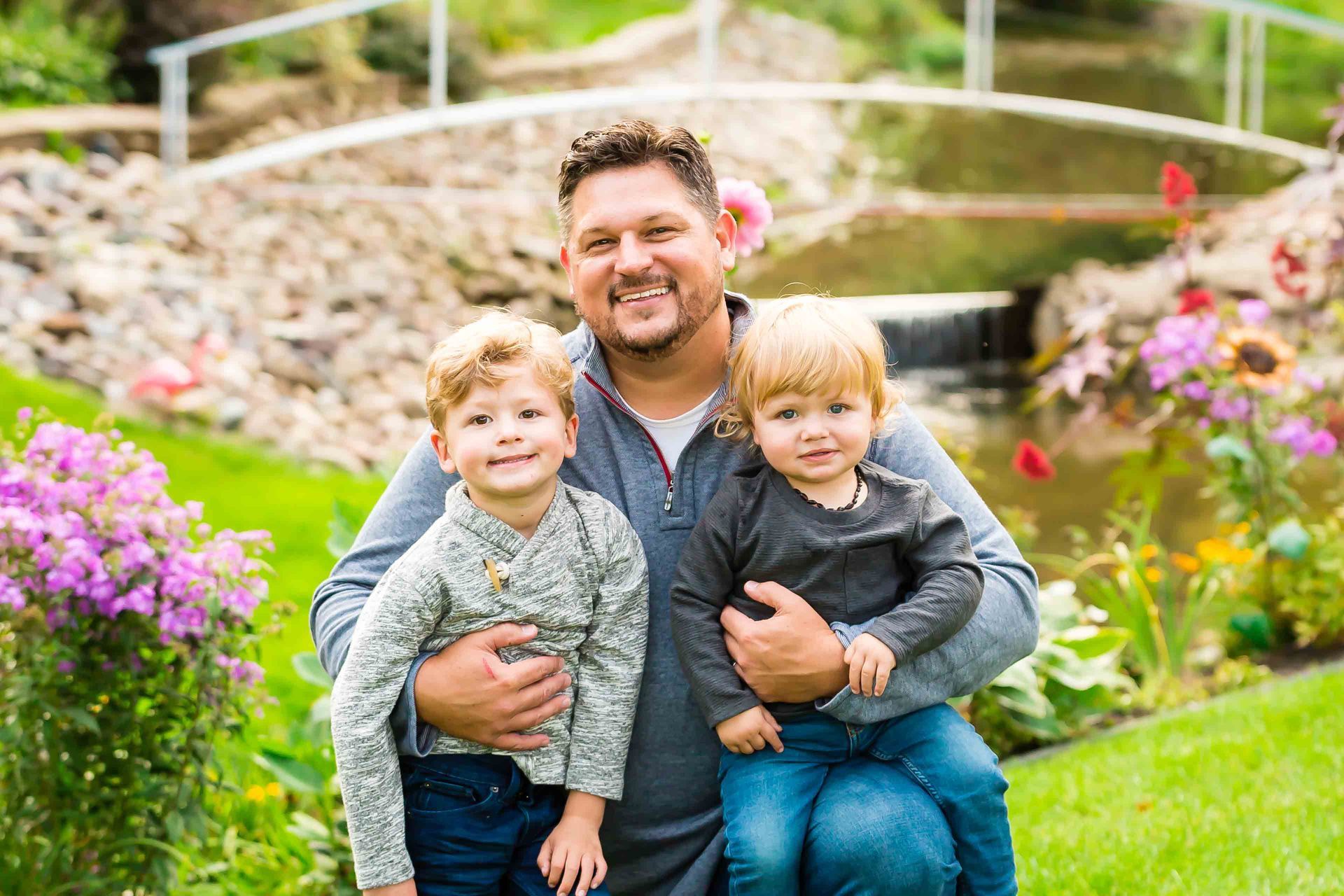 Man holding two young children, smiling in a garden with a bridge and flowers.