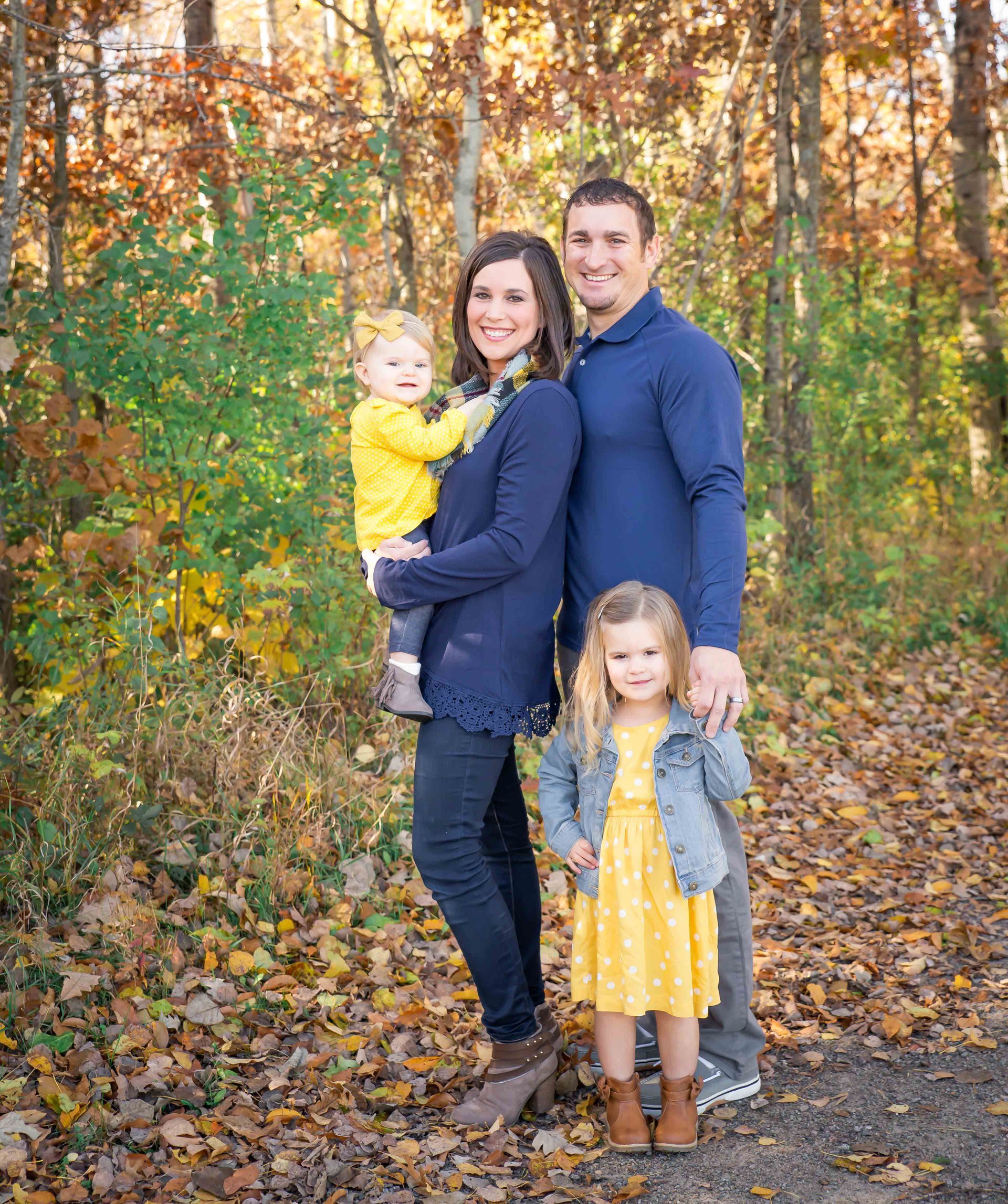 Family of four poses outdoors in fall foliage. Two children in yellow outfits, parents in blue, smiling.