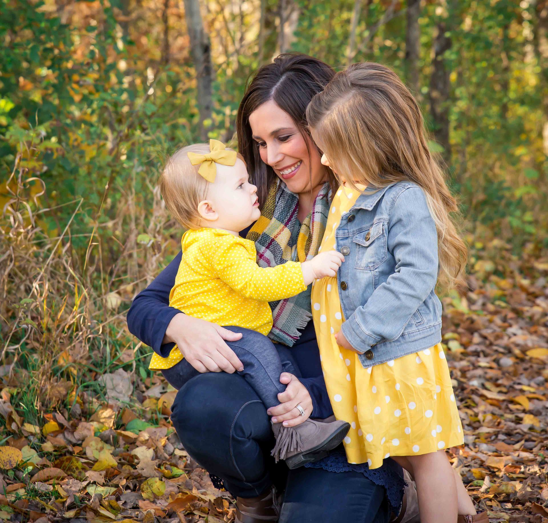 Woman embracing two children in fall foliage. One child in yellow, other in denim jacket.