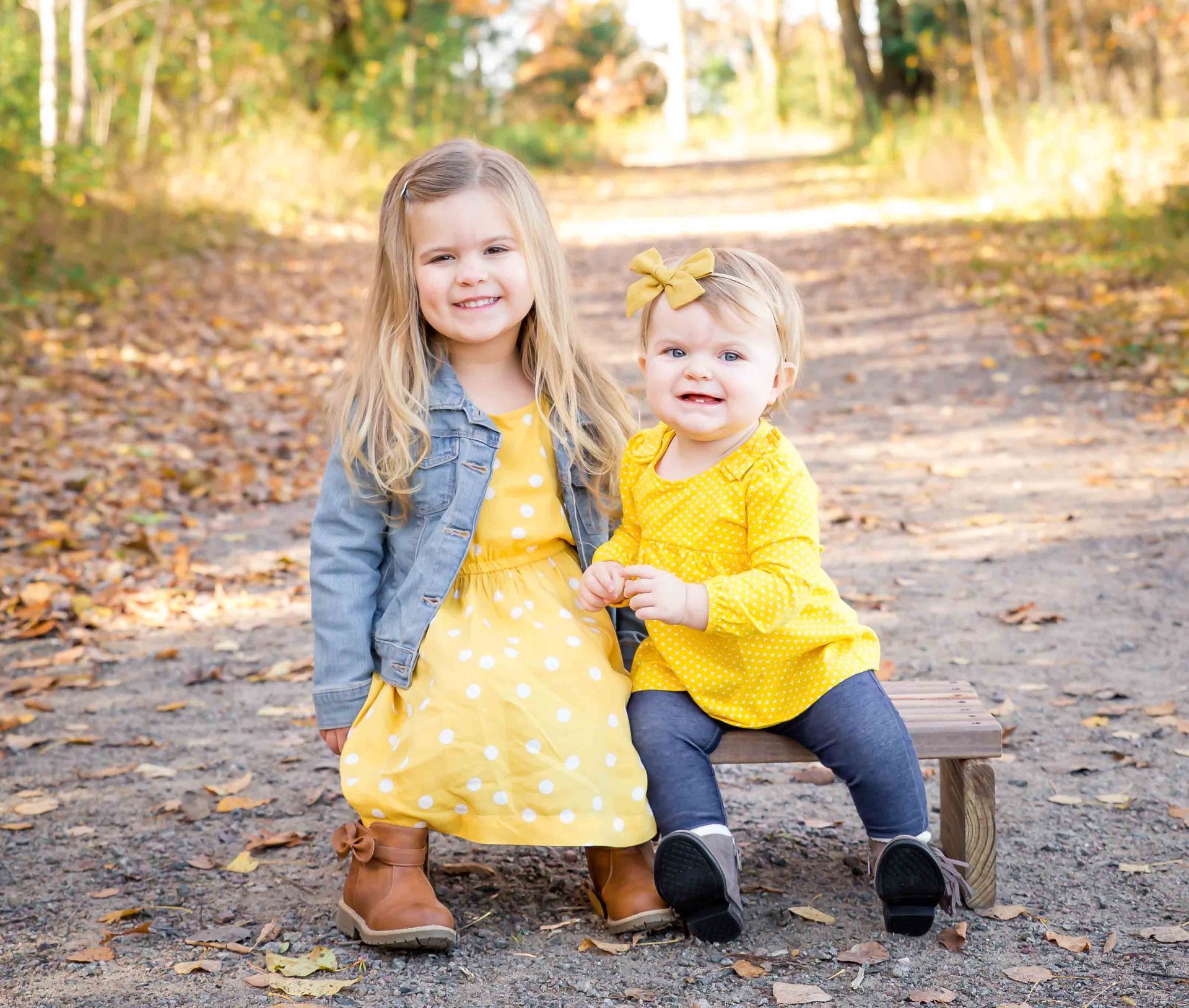 Two young girls smiling outdoors on a path, one in a yellow dress and jacket, the other in yellow top and jeans.