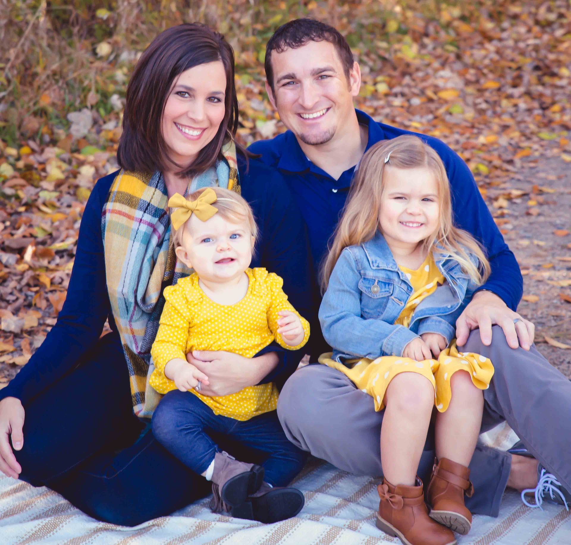 Family of four smiling outdoors, seated on blanket. Fall setting with foliage.