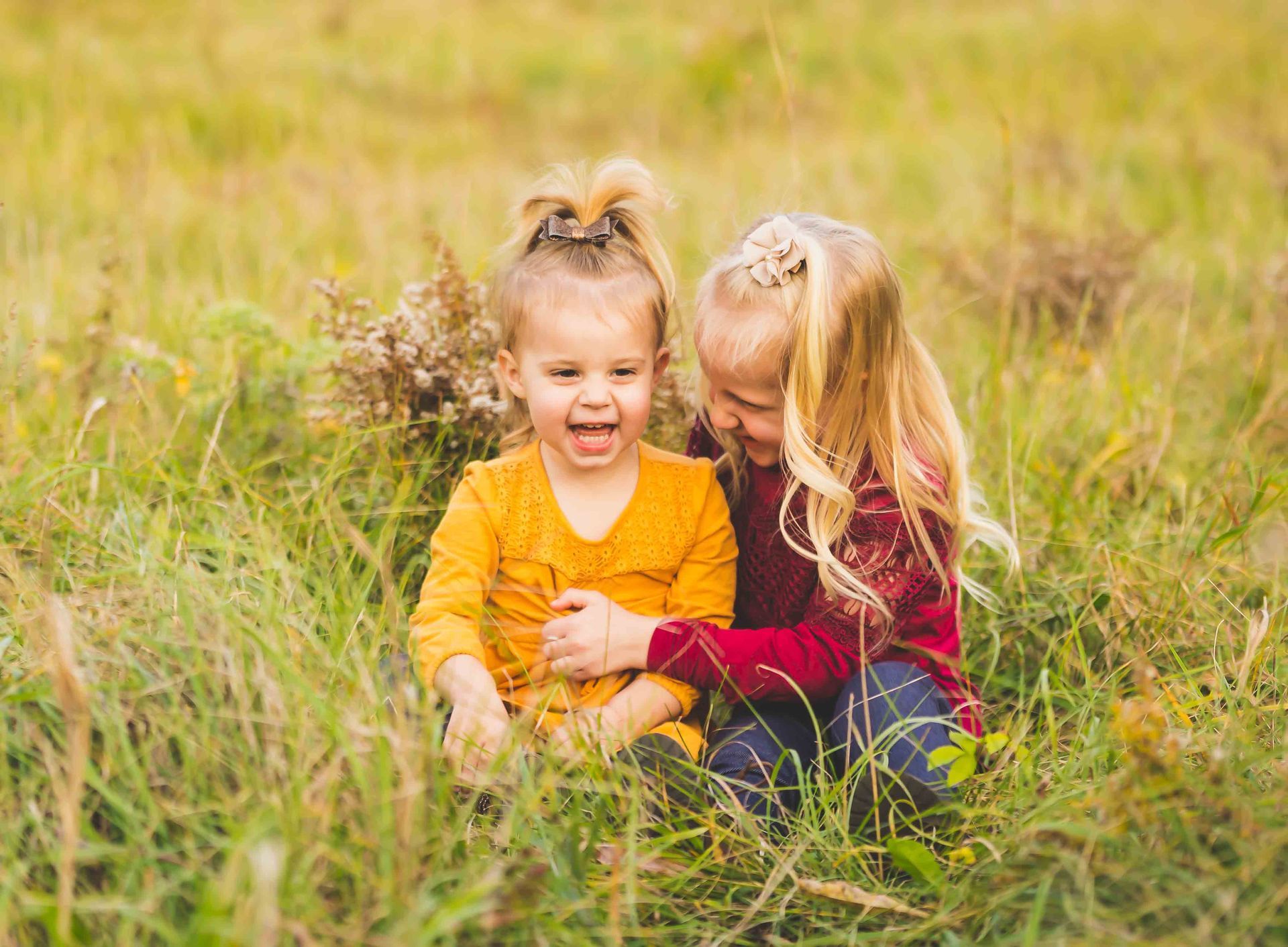 Two young children laughing in a grassy field. One in yellow, the other in red, embracing.