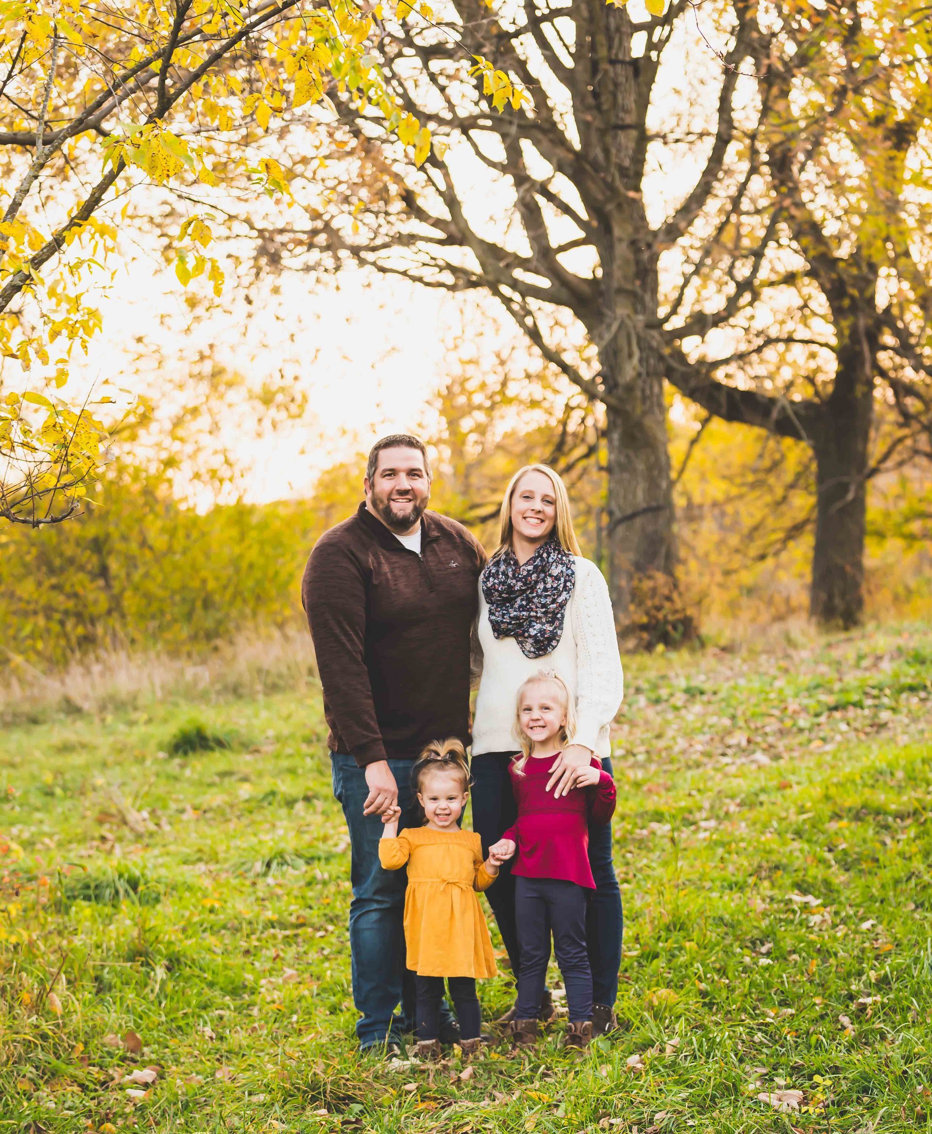 Family of four poses in a park with autumn foliage. Parents smile holding hands with two young children.
