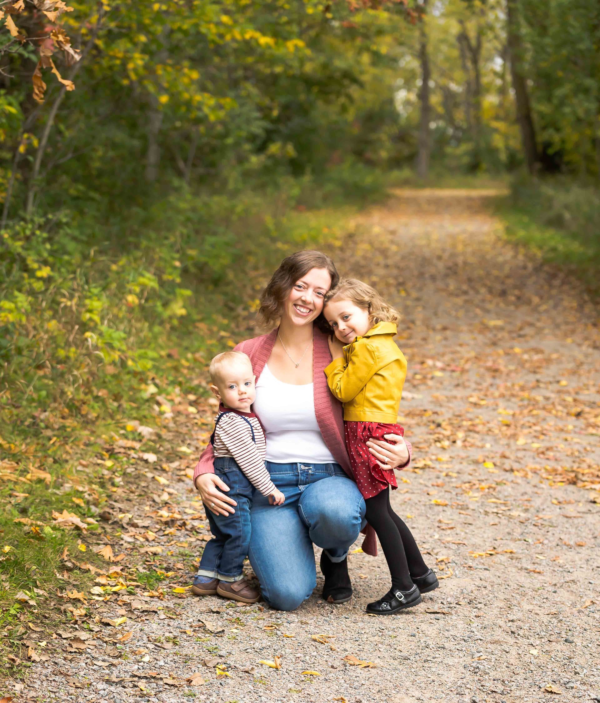 Woman with two children on a wooded path, kneeling and embracing them.