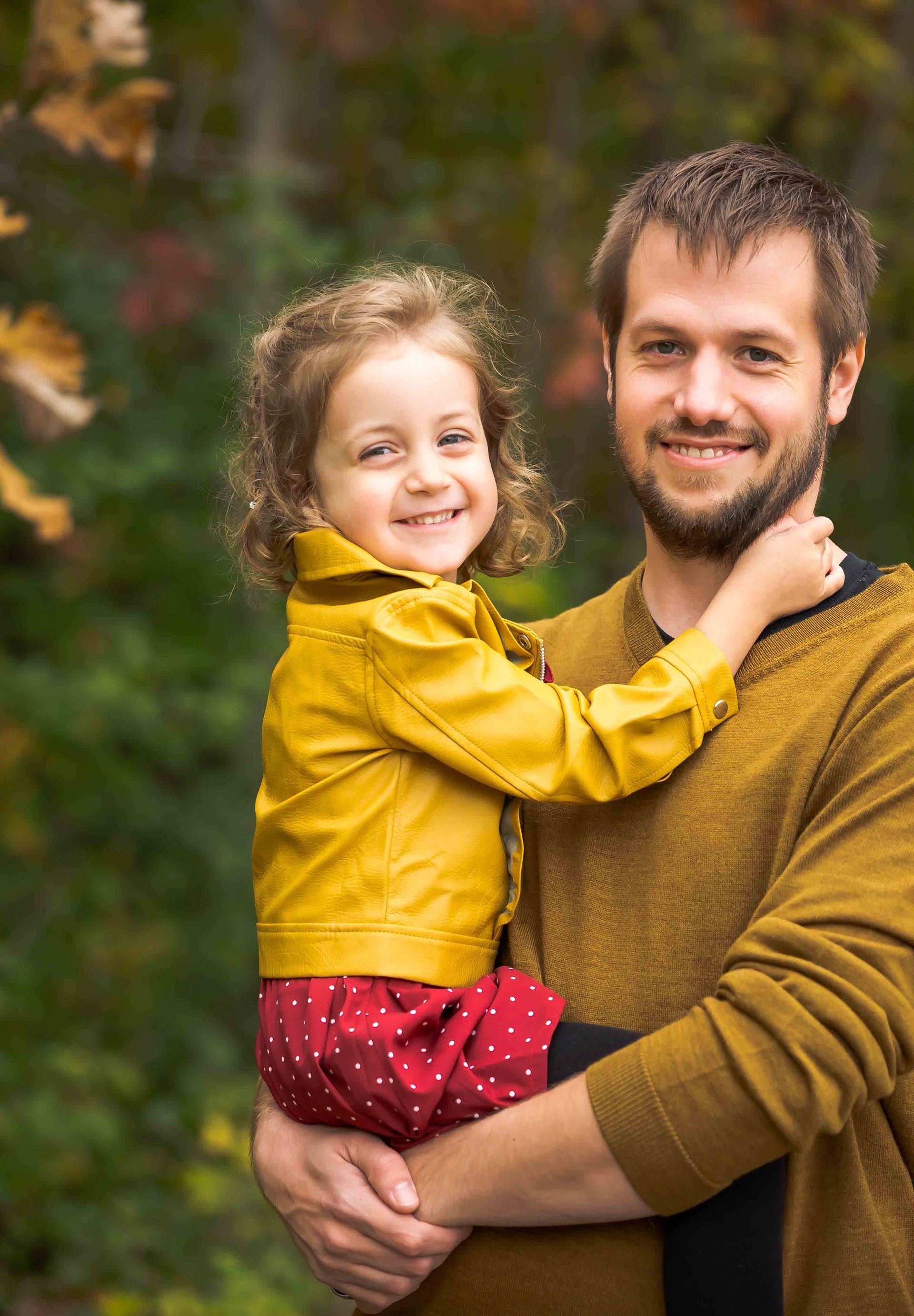 Man holding a smiling child, both wearing yellow and gold. Background of green and autumn leaves.