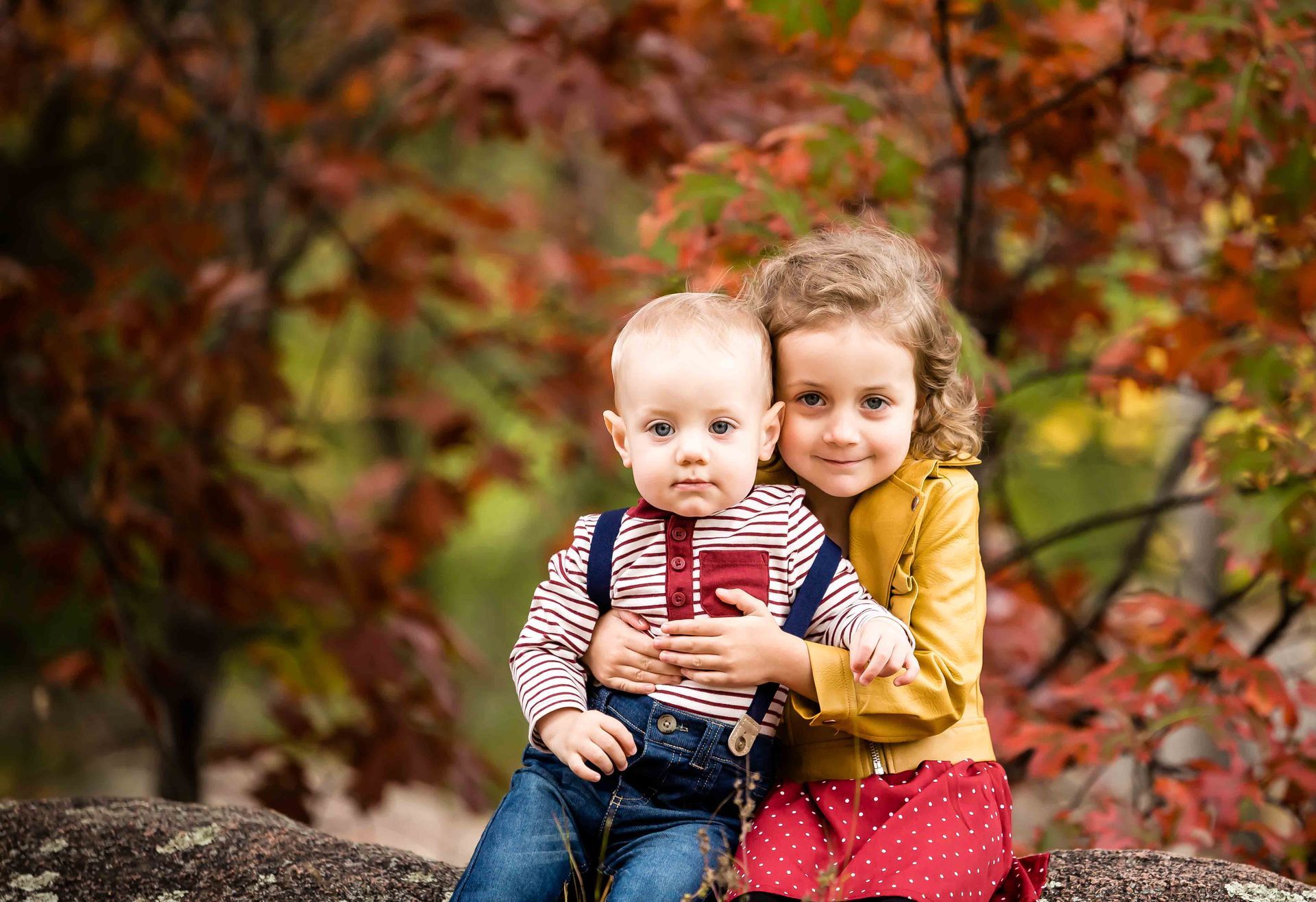 Girl hugs baby, both sitting outdoors. Fall foliage in background.