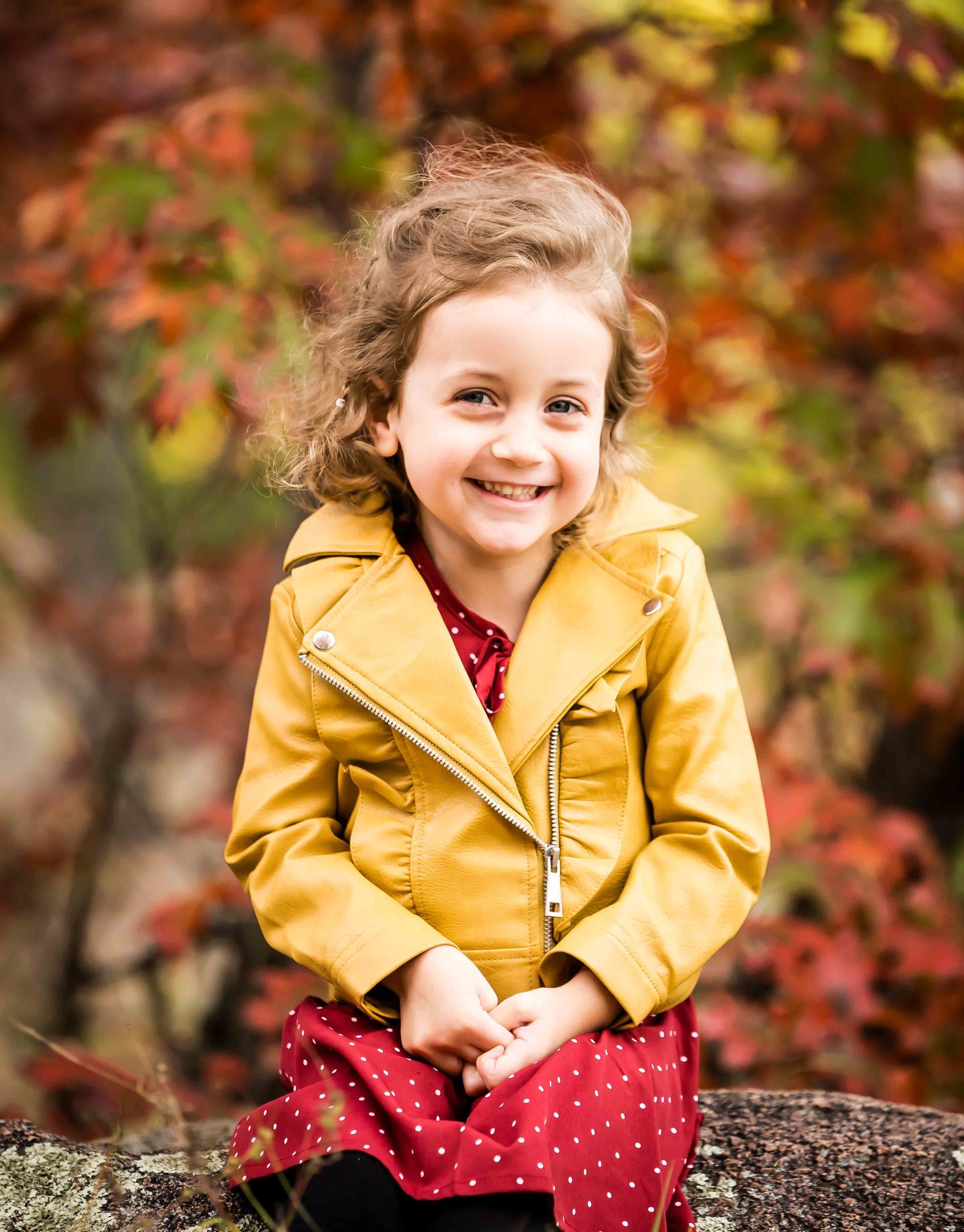 Girl in yellow jacket and red dress smiles outdoors. Autumn foliage in background.