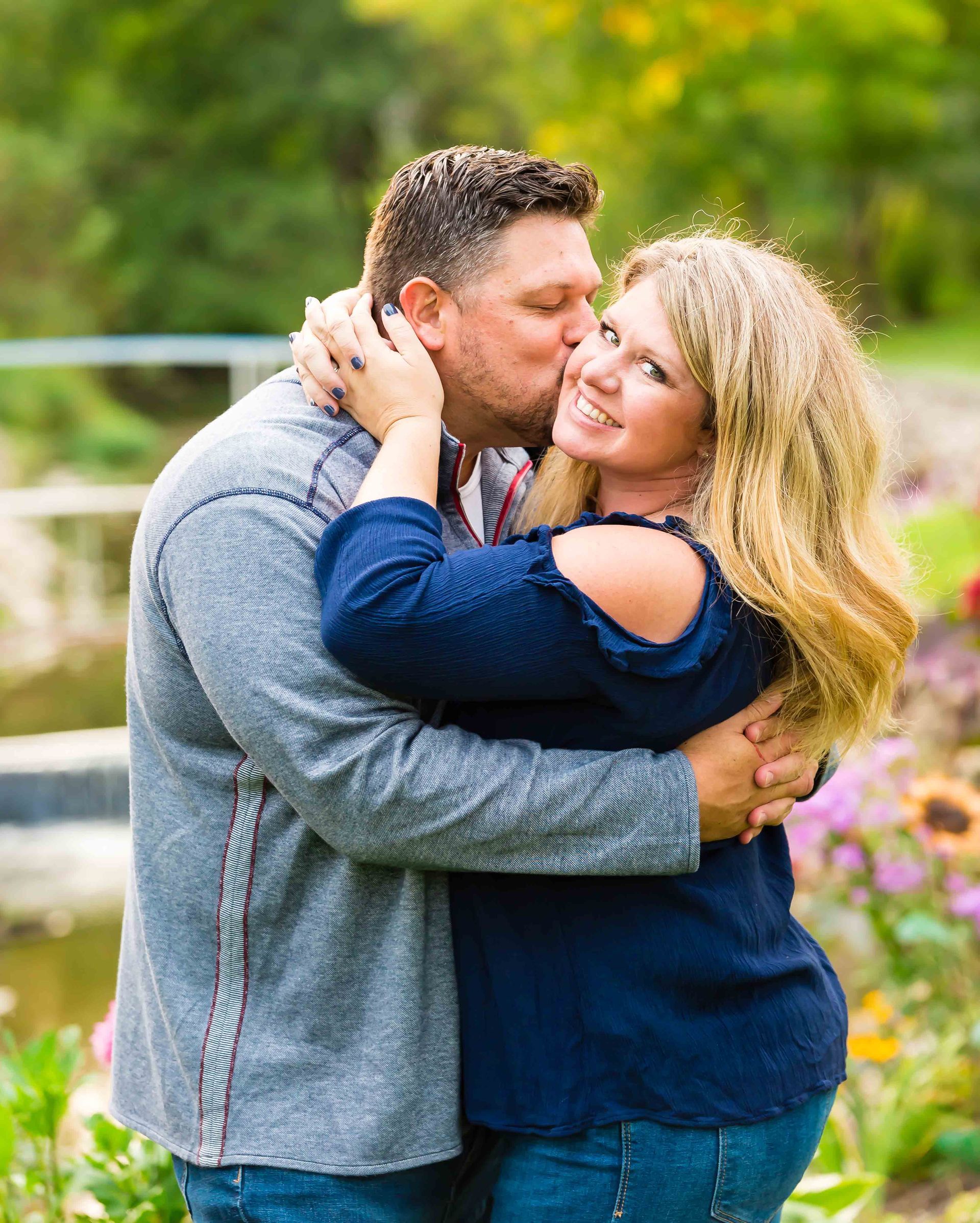 Man kisses woman's cheek in a loving embrace, outdoors. She smiles; he wears a gray shirt. Flowers in background.