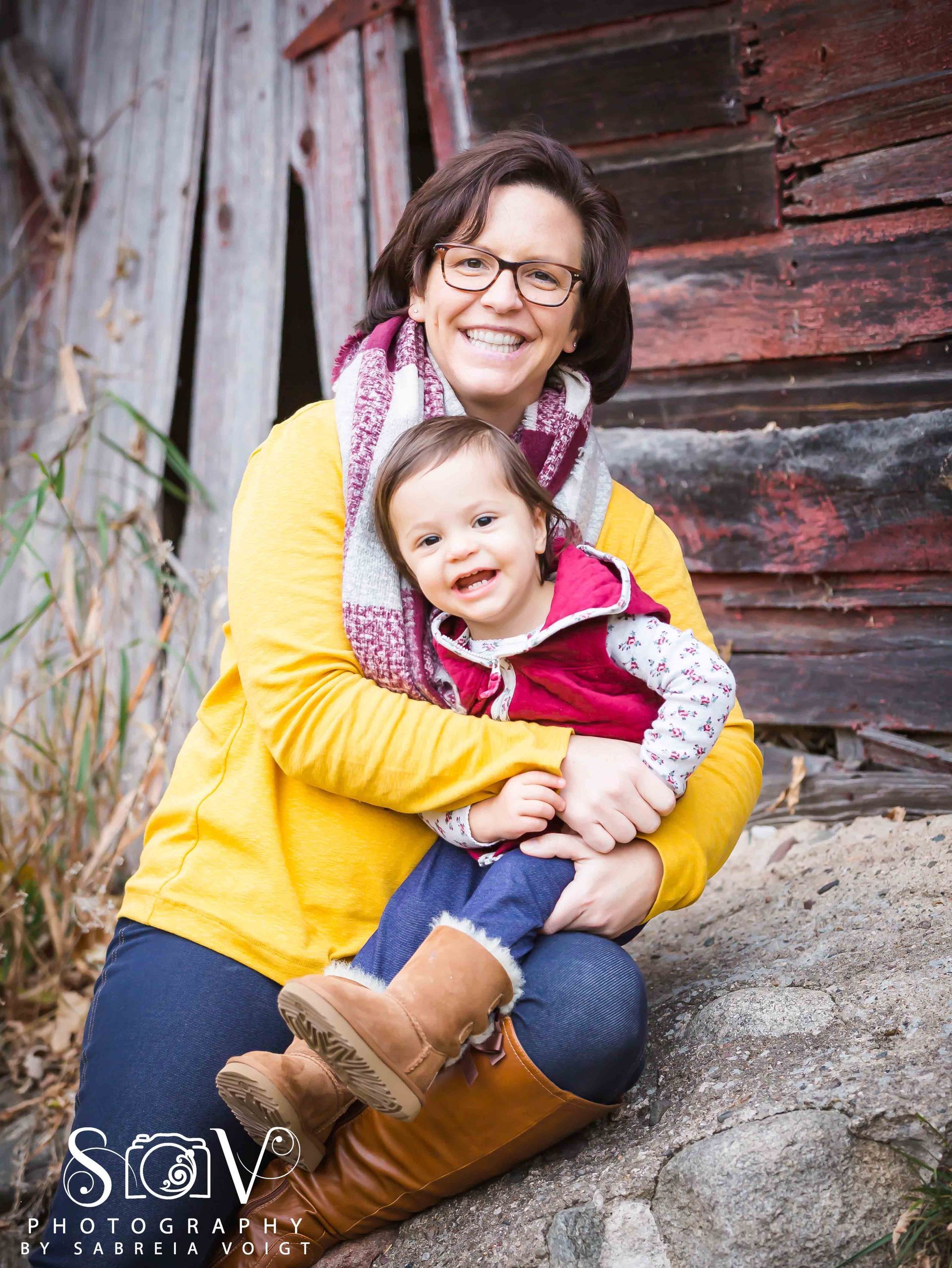 Woman in glasses hugs toddler, both smiling. They are outside near a weathered wooden building.