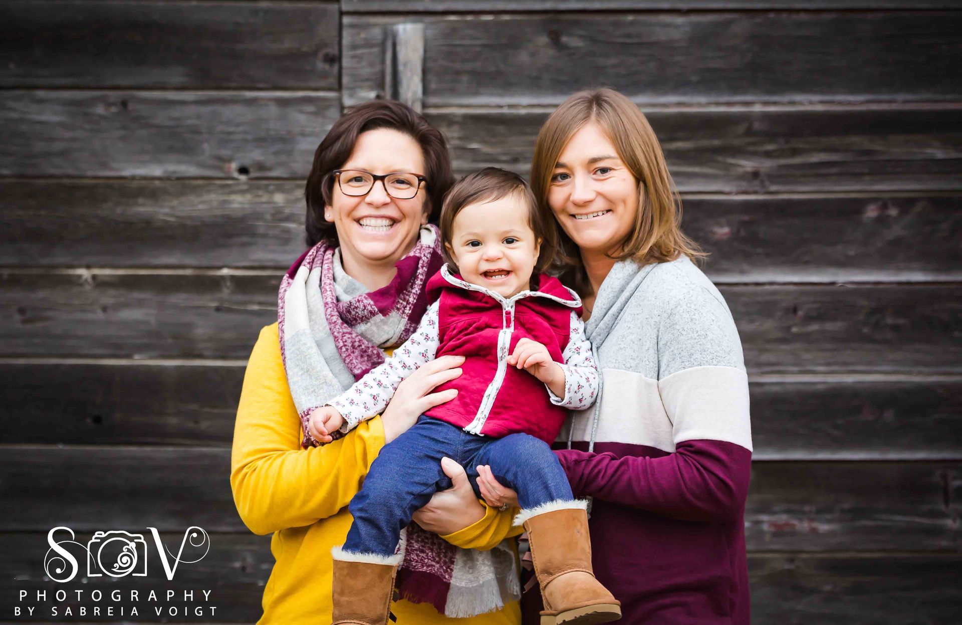 Two adults and a child pose in front of a weathered wooden wall. They smile at the camera.