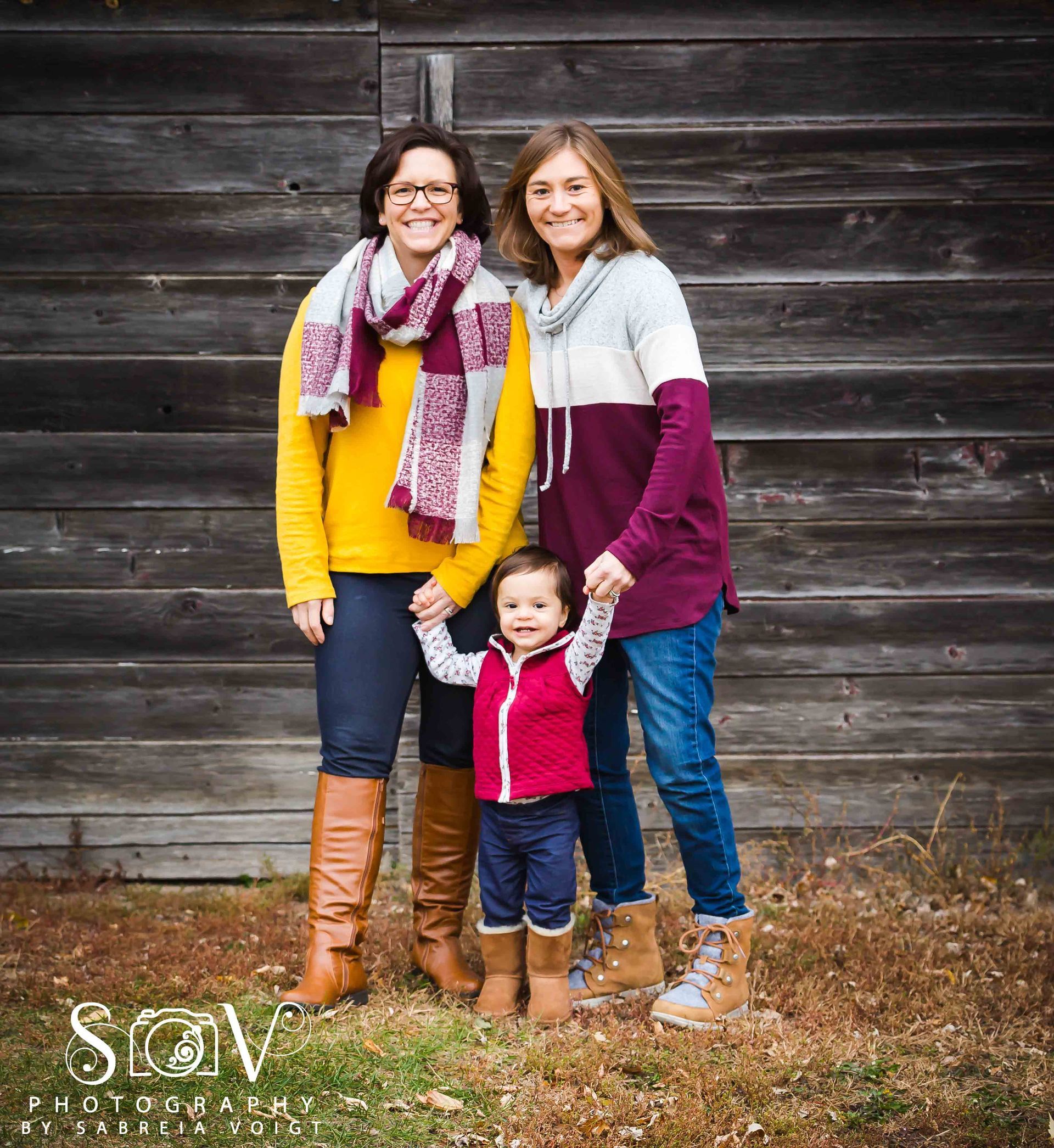 Two women and a toddler holding hands in front of a wood wall; women wearing sweaters and boots; child in a vest.