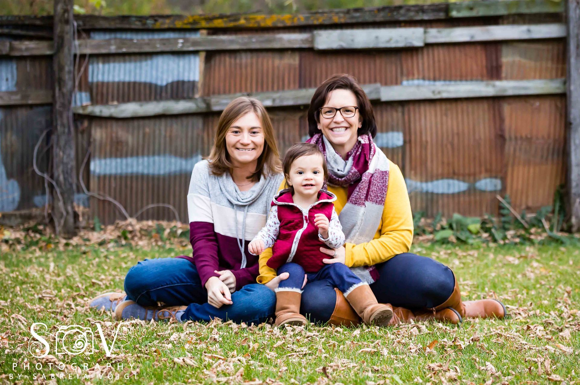 Woman, child, and person with glasses seated on grass in front of a rustic fence; smiling.