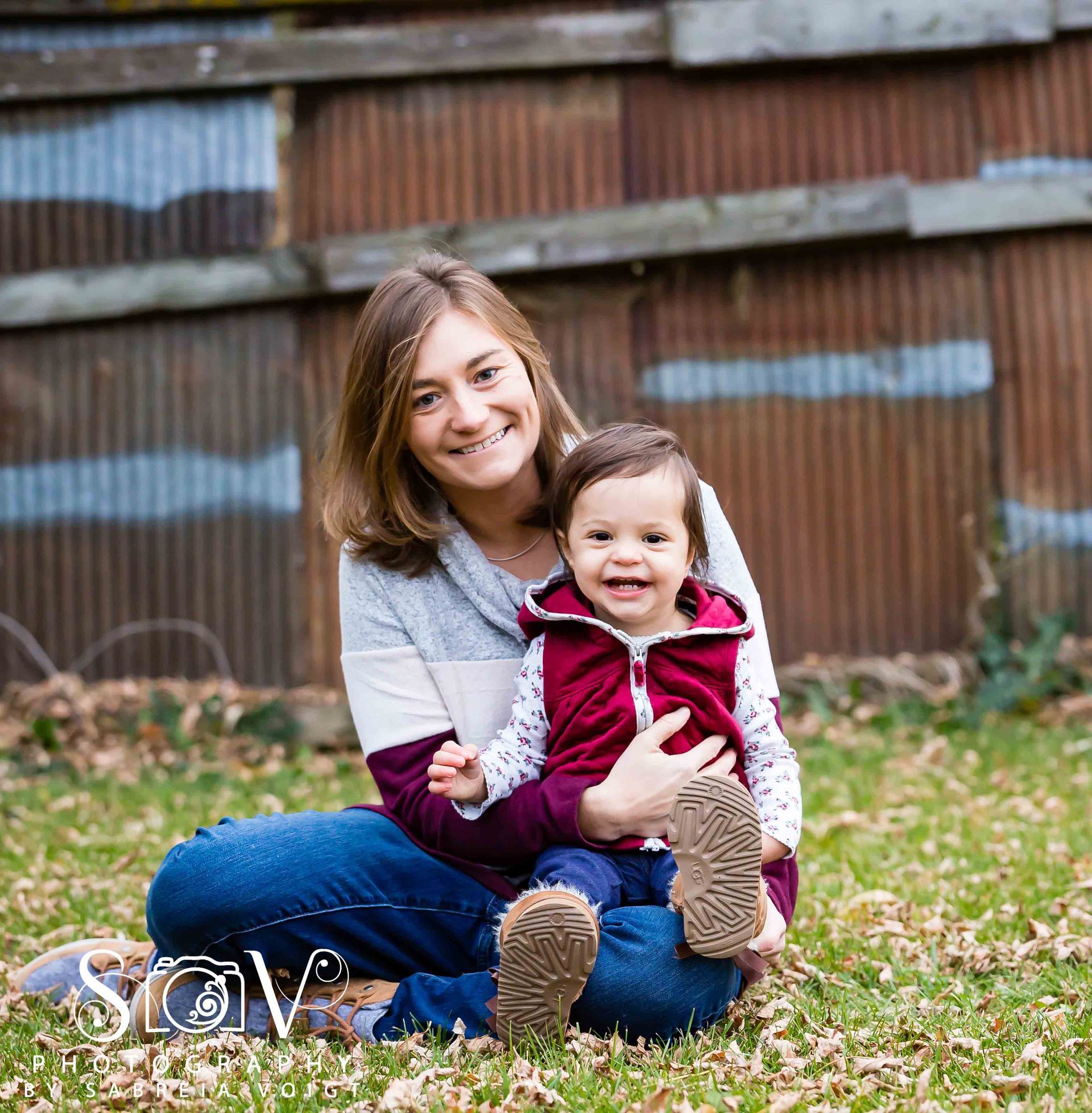 Woman and baby smiling outdoors, sitting on grass; rusted metal background.