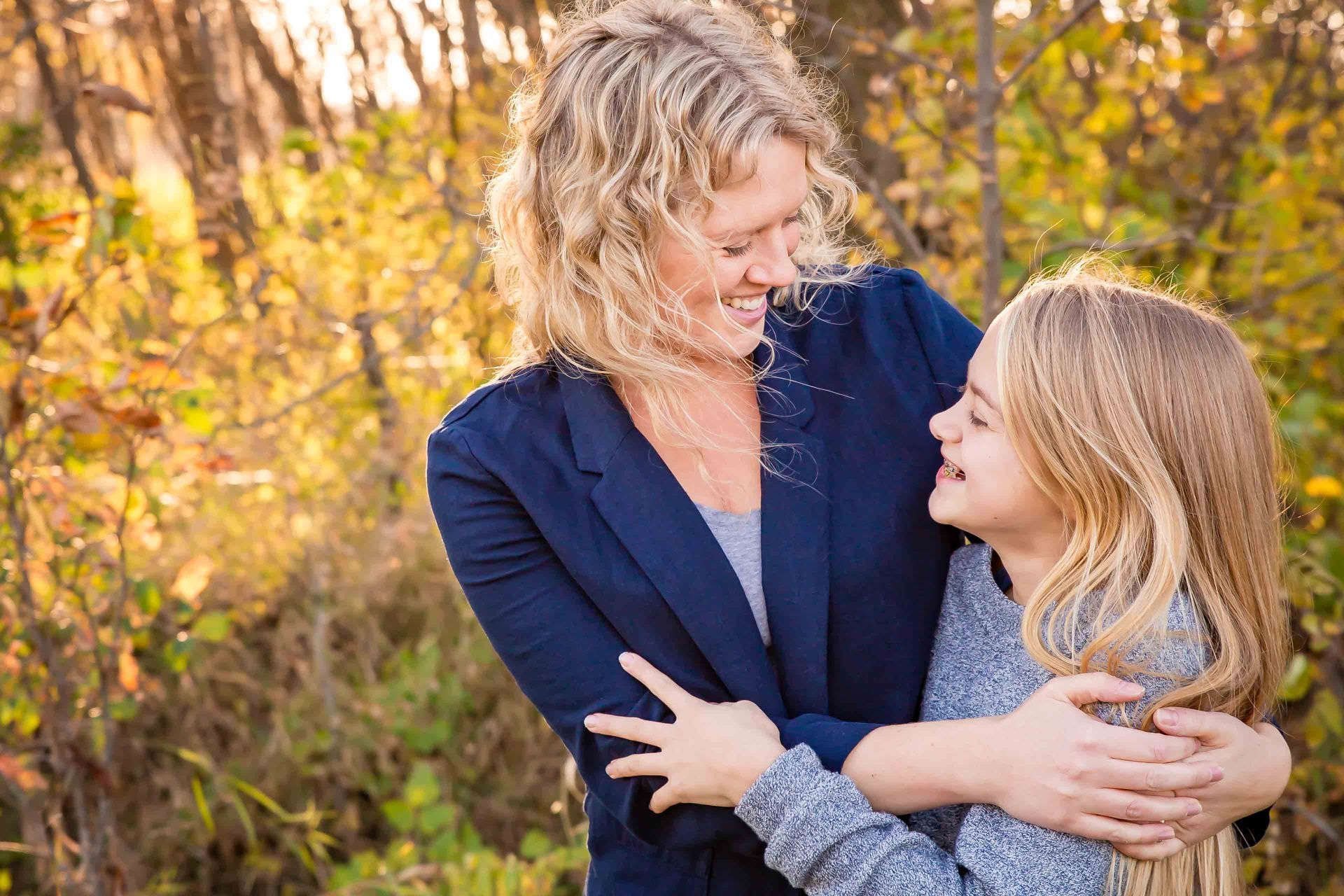 Woman in blue jacket embraces a girl; both smile outdoors in sunlight.