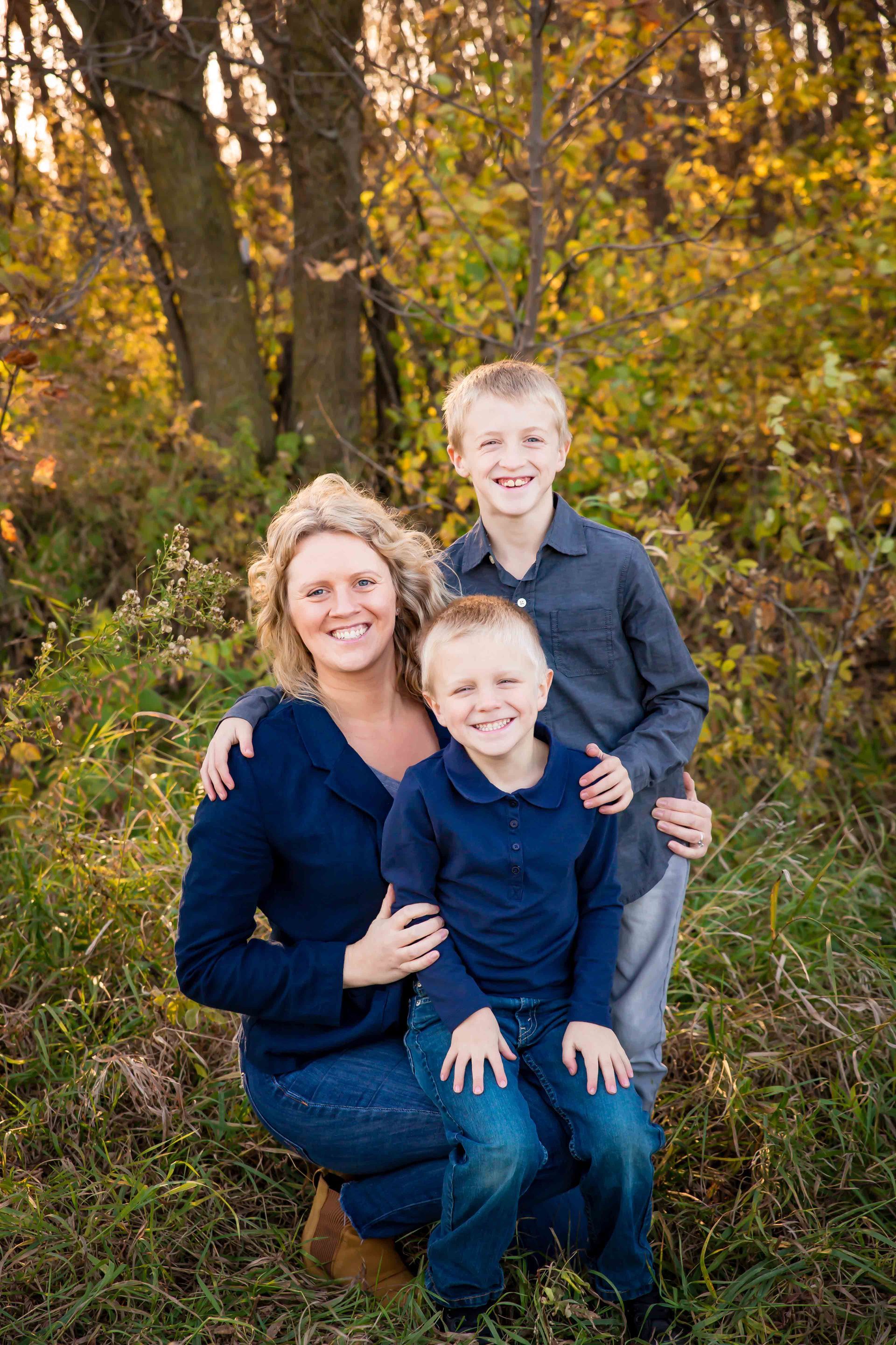 Woman and two children pose outdoors in front of fall foliage; all smile at the camera.
