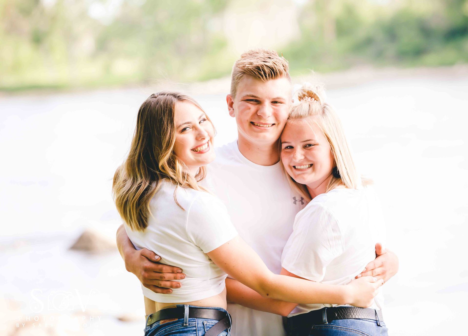 Three people in white shirts embrace outdoors, smiling near water.
