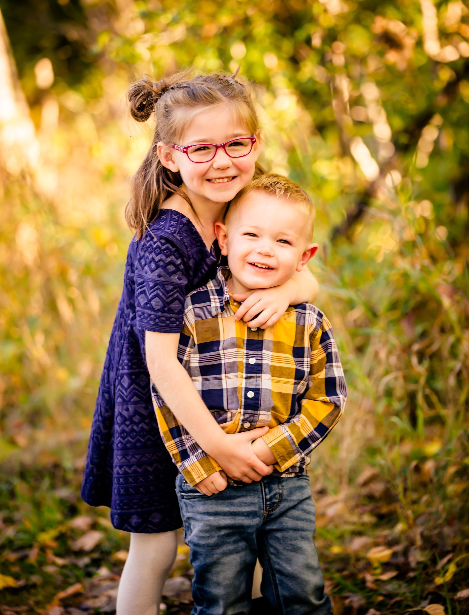 Girl with glasses hugs a smiling boy outdoors, both looking at the camera. Autumn leaves in background.