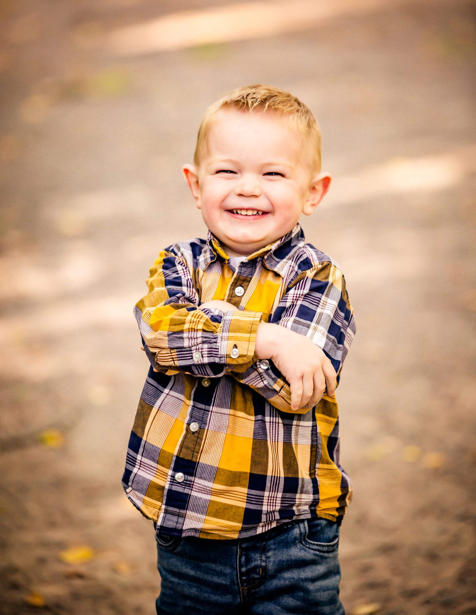 Blond-haired child in plaid shirt smiles, arms crossed, standing on a path.