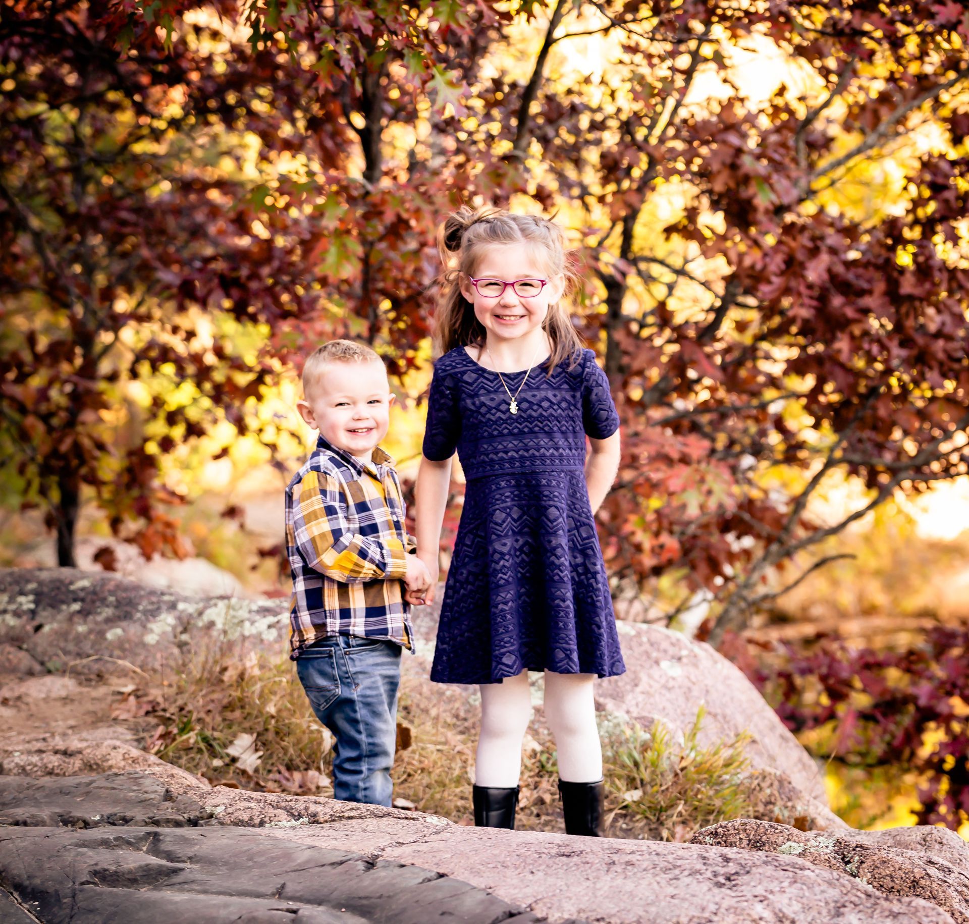 Two children smiling, holding hands, standing on rocks, autumn foliage background.