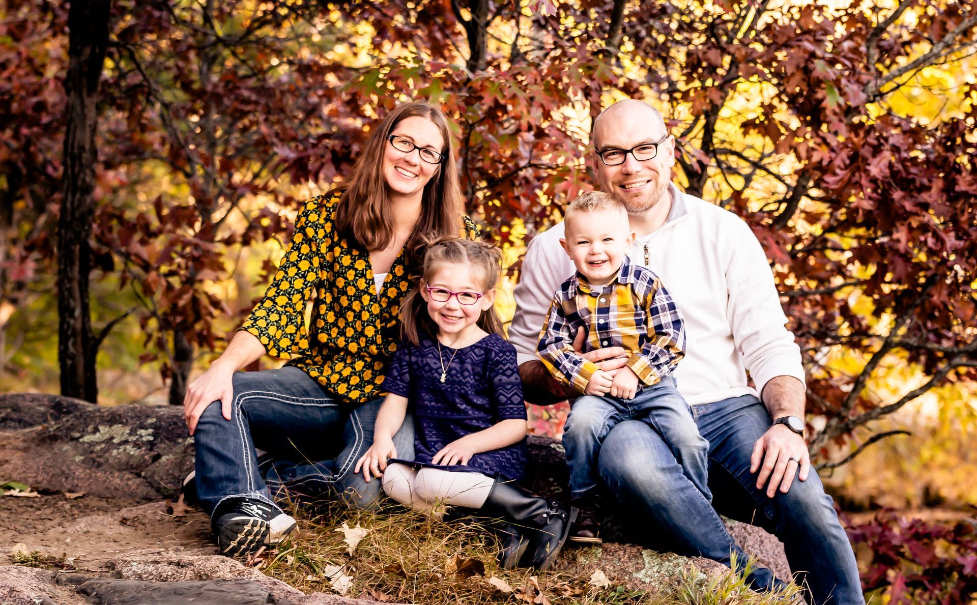 Family of four sits on rocks with autumn foliage. Smiling, wearing casual clothes.