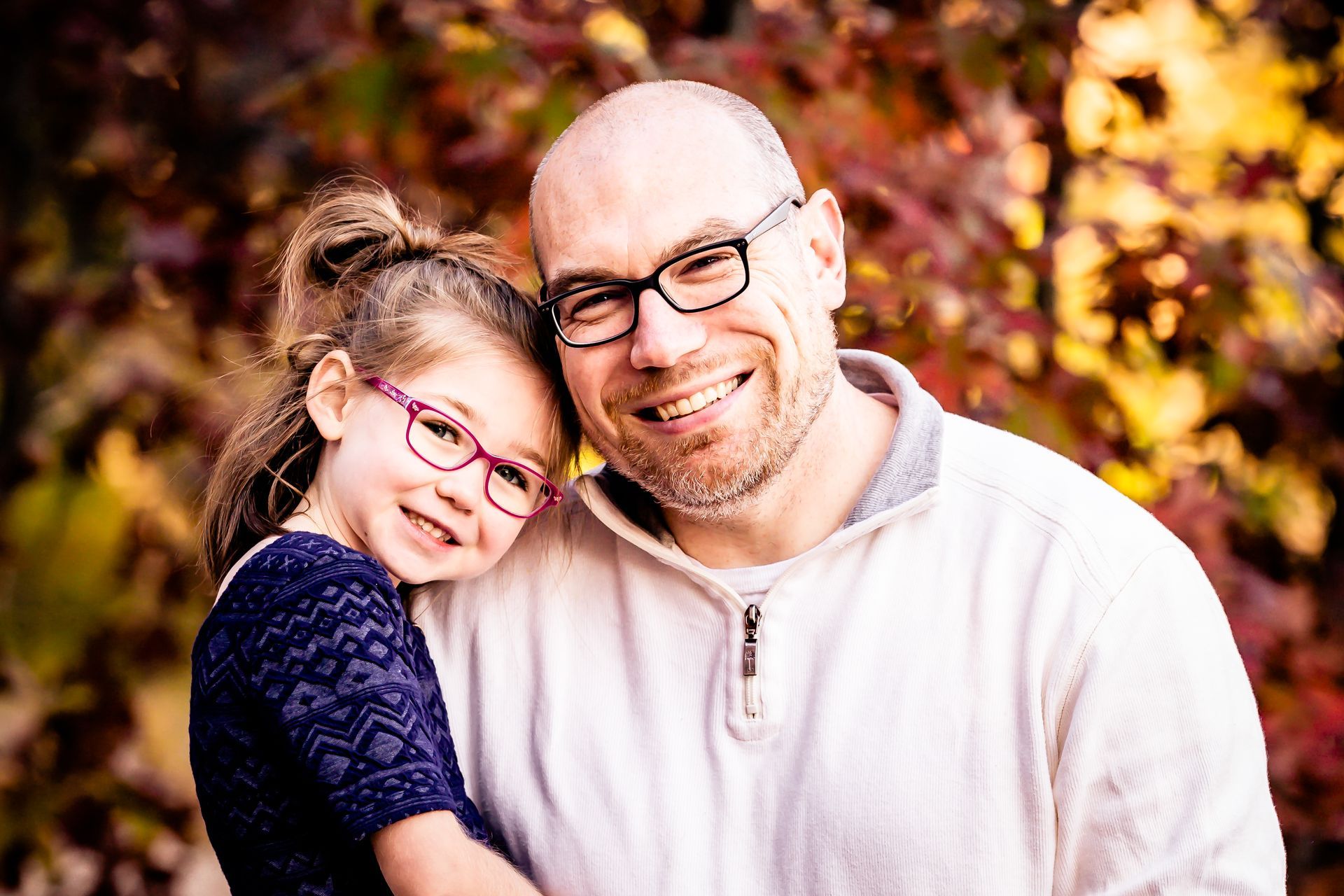 Father and daughter in glasses smiling; autumn foliage in the background.