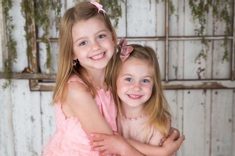 Two young girls in pink dresses smile and hug, in front of a rustic white wooden backdrop.