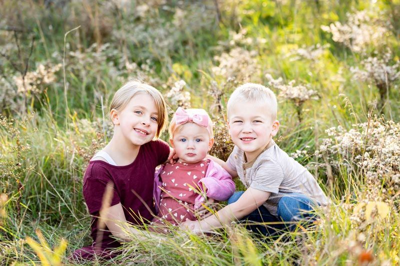 Three children smiling, sitting in tall grassy field.