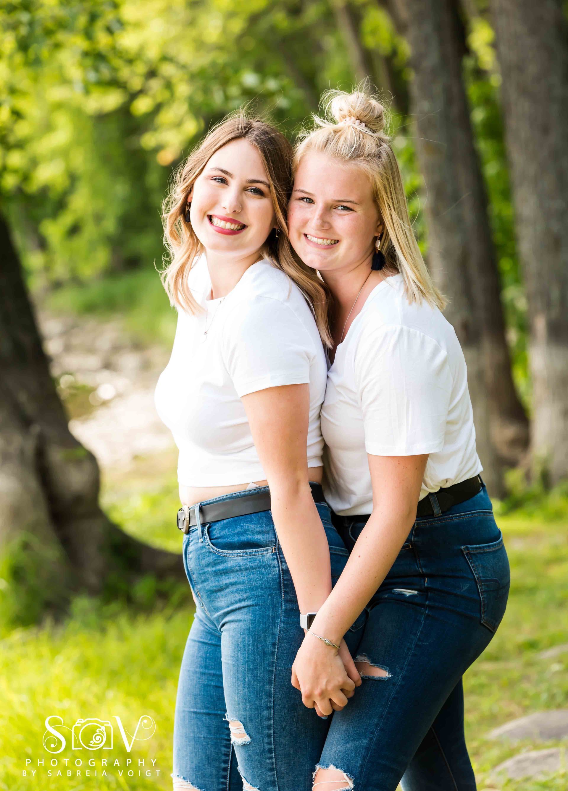 Two women smiling, holding hands, wearing white shirts and jeans, posing outside.