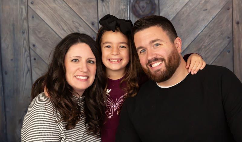 Family of three smiles at the camera against a wooden door backdrop.