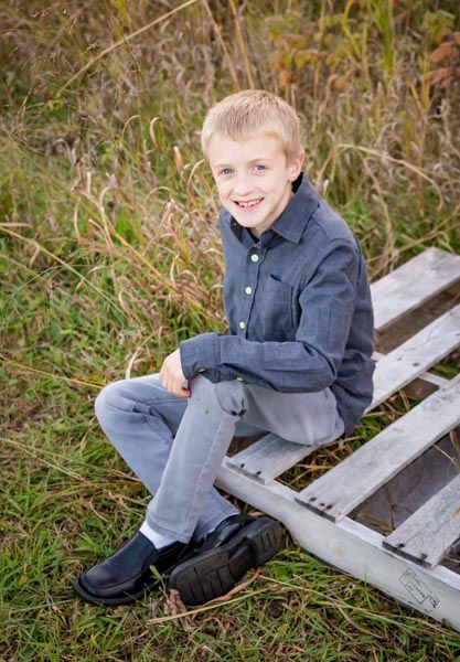 Boy sitting on a wooden pallet outdoors, smiling. He wears a dark blue shirt, gray pants, and black shoes.