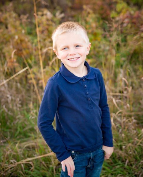 Blond-haired boy smiling, wearing a navy blue collared shirt and jeans, standing in a field.