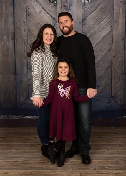Family of three smiling, posing in front of a wooden backdrop. Child in burgundy dress, parents in casual clothing.