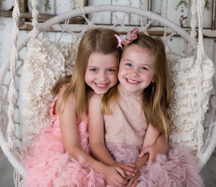Two young girls smiling in a hanging chair. One in pink, the other in light pink.
