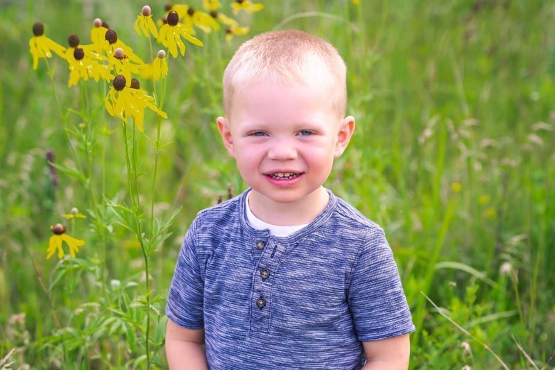 Young child smiling in a field of green grass and yellow flowers; wearing a blue shirt.