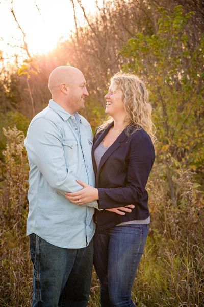 Couple smiles at each other, outdoors. Man in blue shirt, woman in blazer and jeans. Sunlight and foliage in background.
