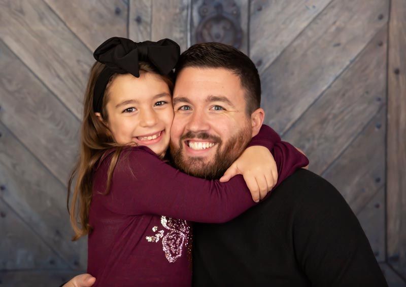 Girl with black bow hugging smiling man against wood background.