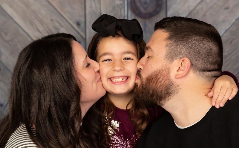 Woman and man kissing a smiling child on the cheeks, against a wood-paneled background.