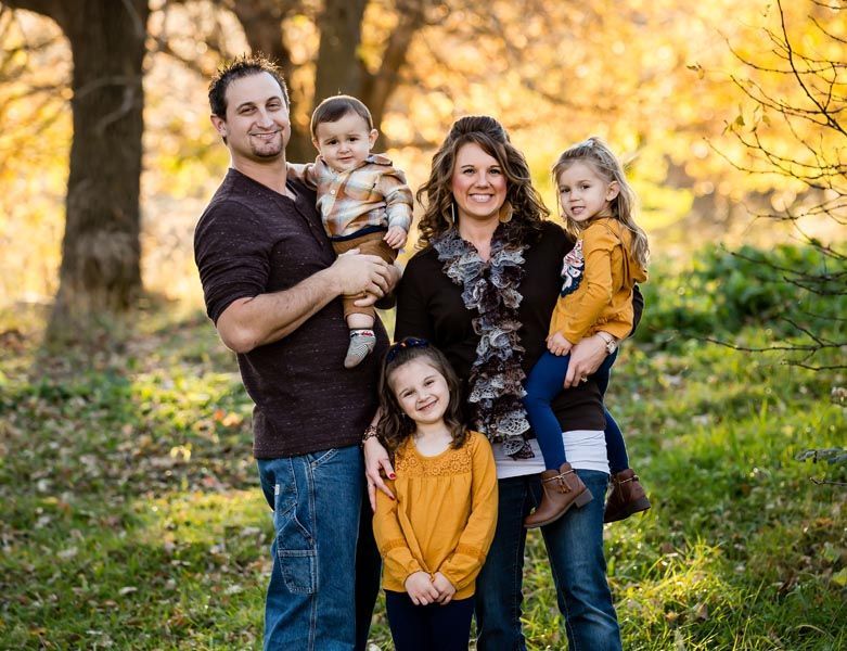 Family of five smiles in an outdoor setting with autumn colors; woman holds a child, man holds a child.