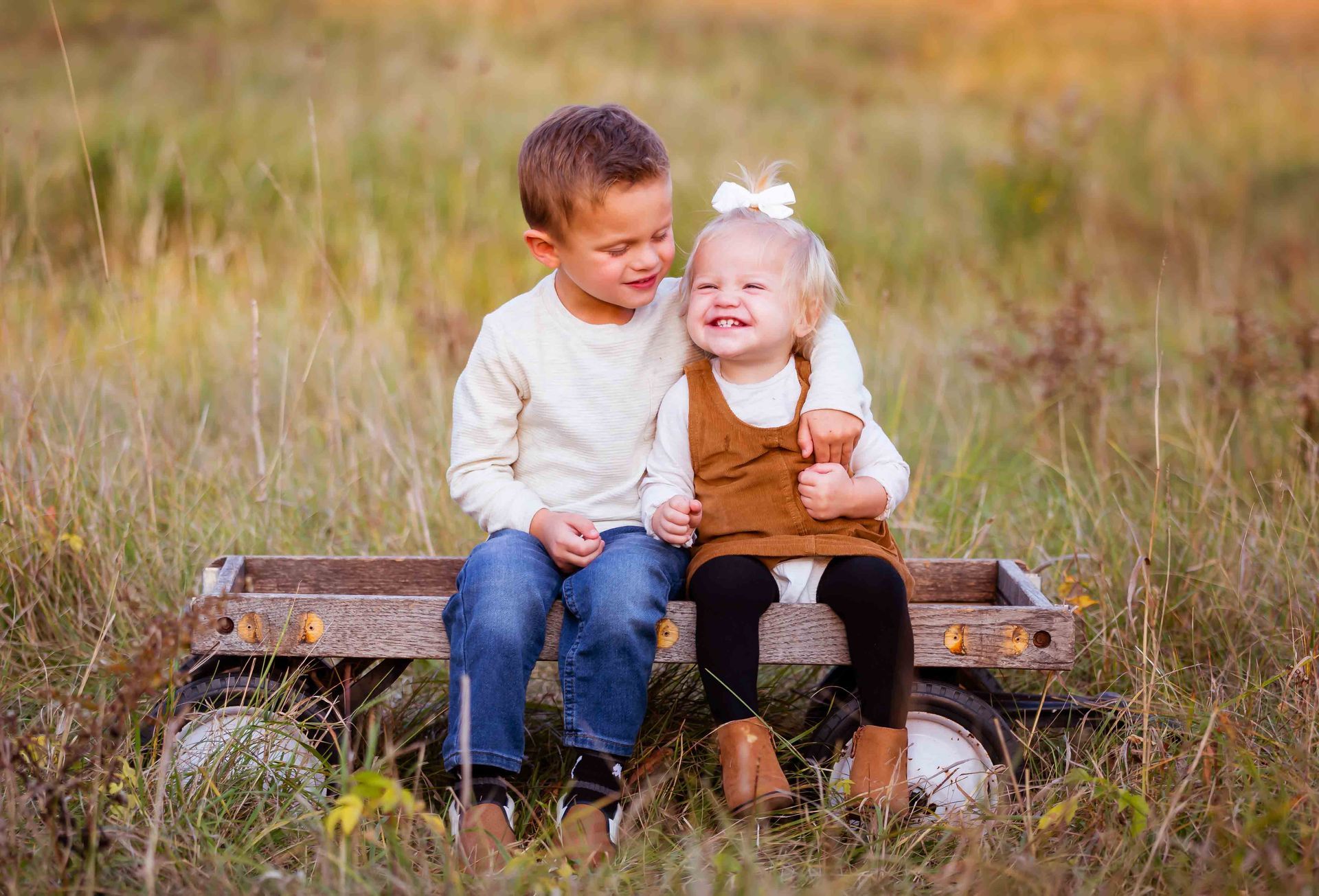 Boy and girl smiling, sitting in a wagon outdoors, boy has arm around girl, tall grass background.