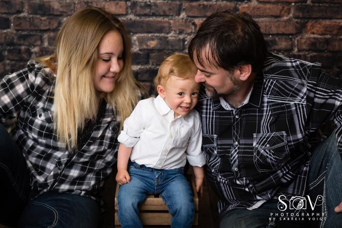Family of three smiles together in front of a brick wall. They are wearing plaid shirts and jeans.