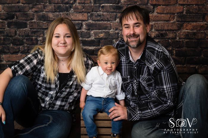 Family of three posing together, sitting in front of a brick wall. All are smiling.
